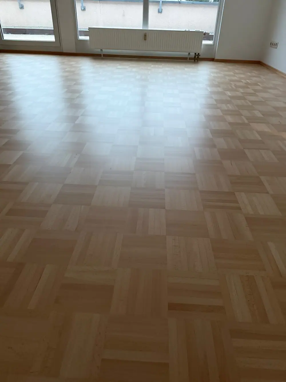 Heller Raum mit abgeschliffenem und matt versiegeltem Mosaikparkett aus Buche vor großen Fenstern und einem Heizkörper.