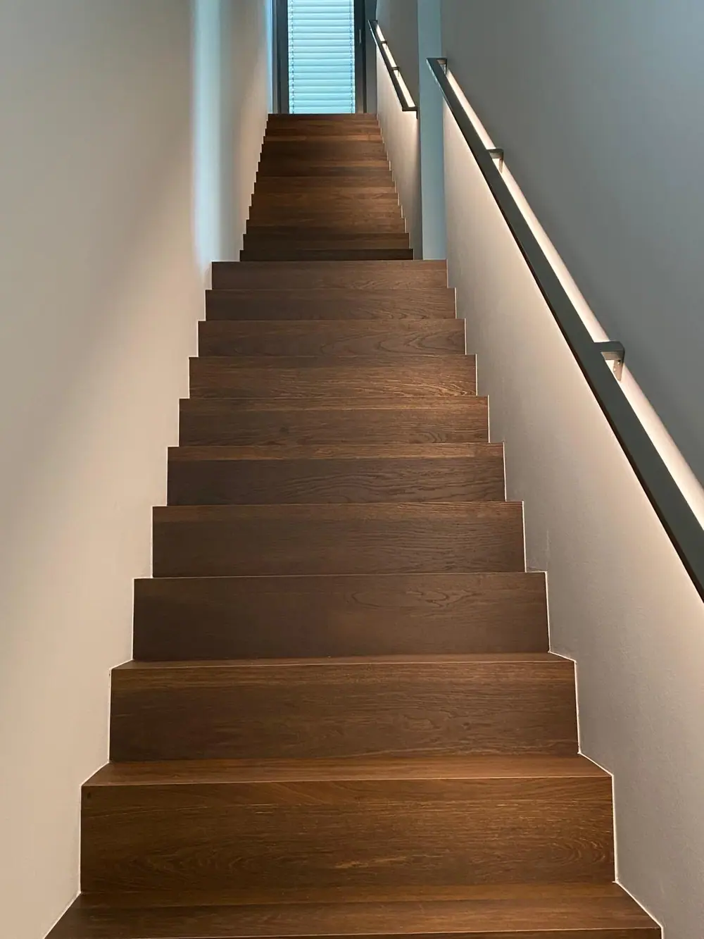 Blick von unten auf einen geraden Treppenlauf mit dunkelbraunen Holzstufen, hellen Wänden und dunklem Handlauf.