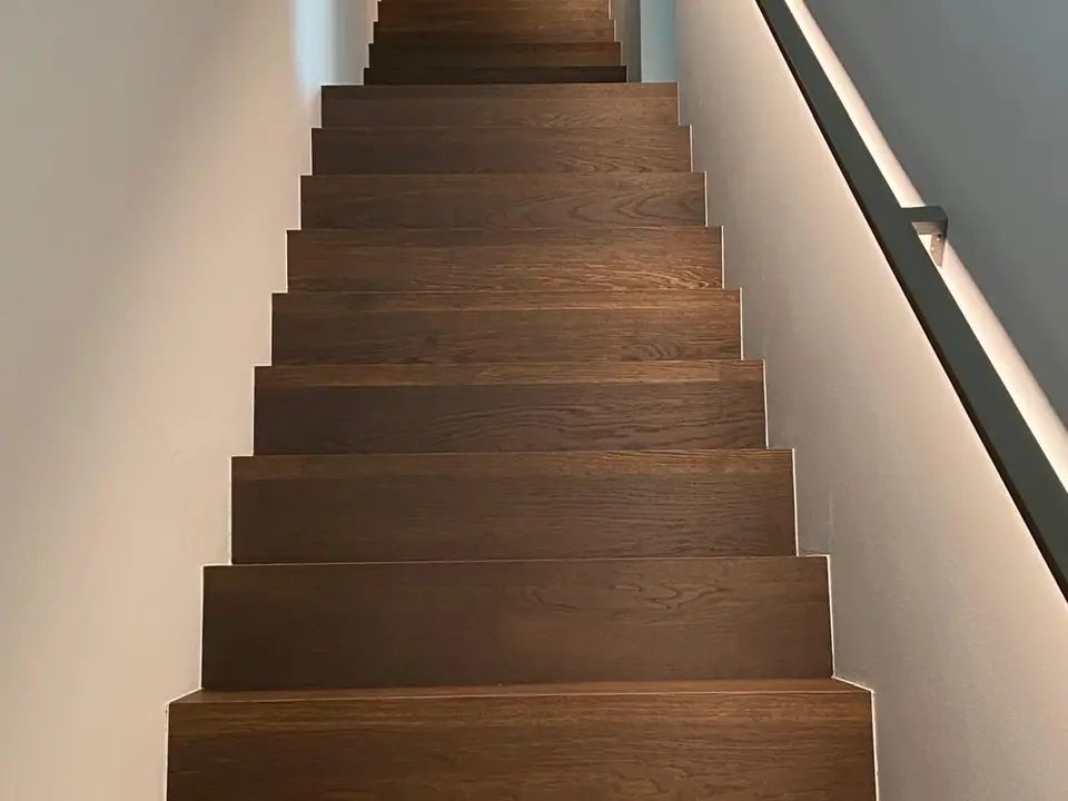 Blick von unten auf einen geraden Treppenlauf mit dunkelbraunen Holzstufen, hellen Wänden und dunklem Handlauf.