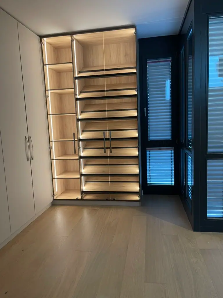 Heller Dielenboden in einem Raum mit weißem Einbauschrank, beleuchtetem Holzregal und dunklen Fensterrahmen.