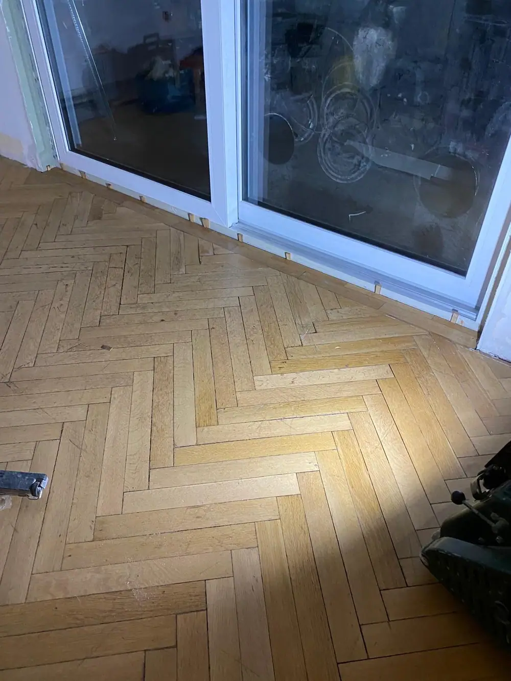 Fischgrätparkett mit geöffnetem Randbereich vor einem Fensterelement.