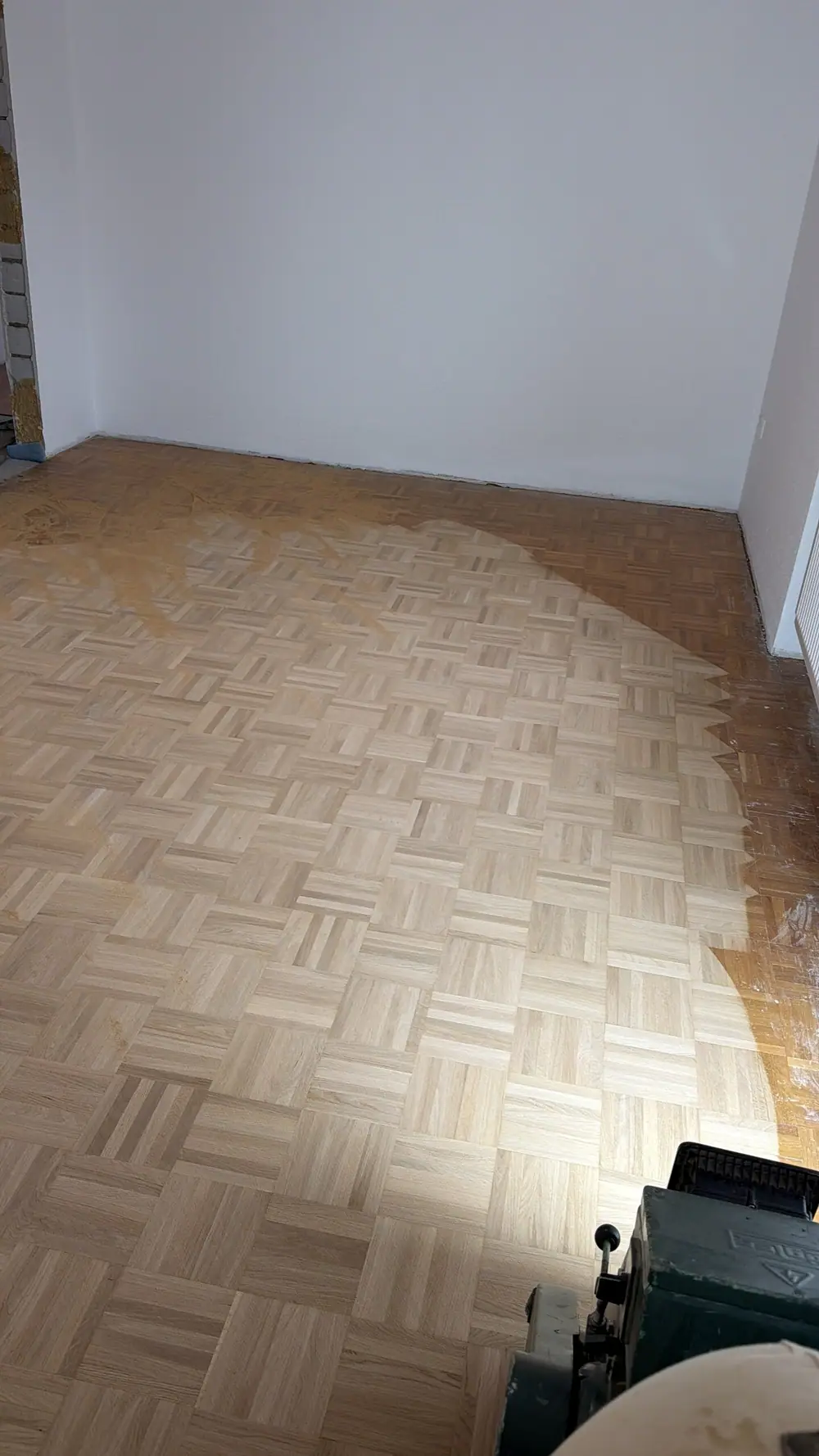 Teilweise bearbeitetes Eiche-Mosaikparkett in einem leeren Raum; die helle bearbeitete Fläche trifft auf den dunkleren Altbestand.