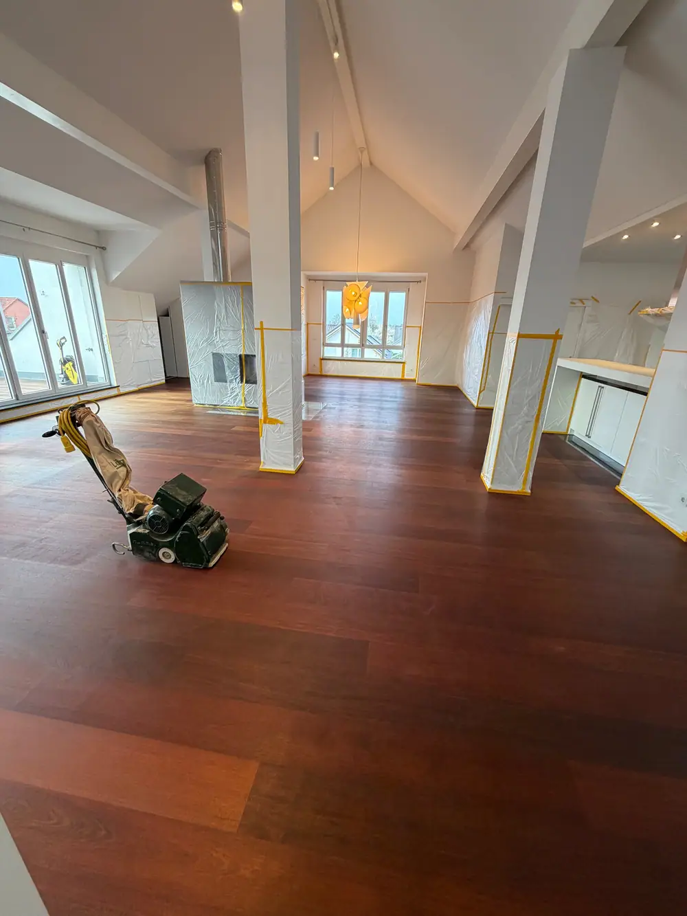 Offener Wohnbereich mit dunklem Landhausdielenparkett, abgedeckten Einbauten und einer Schleifmaschine im Vordergrund.