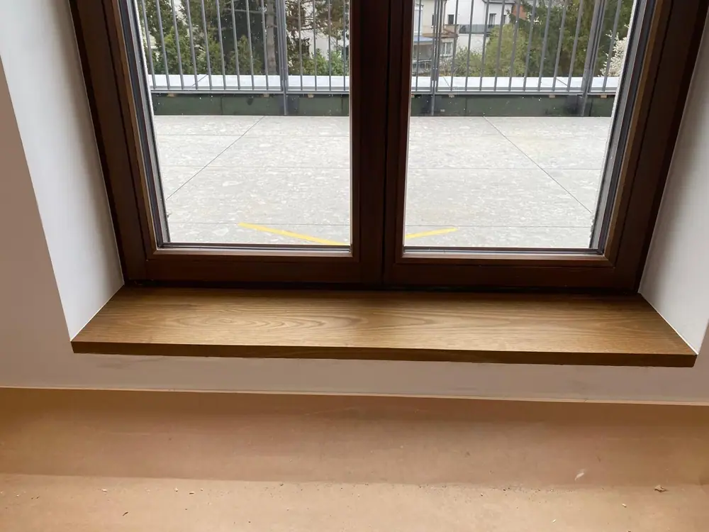 Fensterbank aus mittelbrauner Räuchereiche vor einem zweiflügeligen Fenster mit Balkonblick.