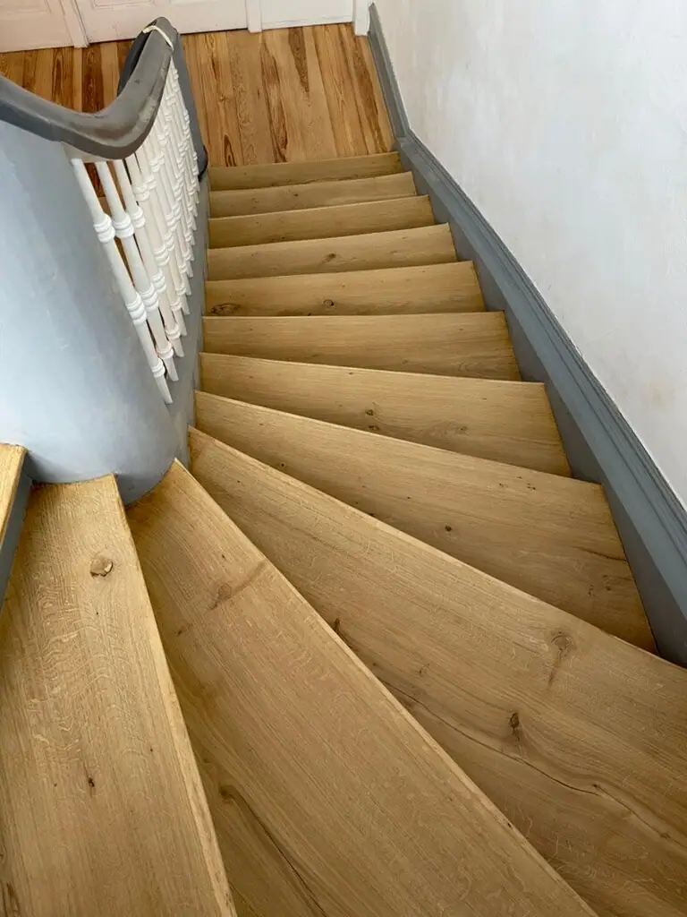 Blick von oben auf eine gewendelte Holztreppe mit hellen Stufen, grauen Wangen und weißem Geländer.