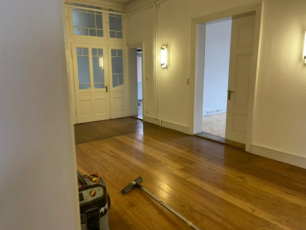 Blick durch einen Flur mit langen Holzdielen, weißen Türen, Wandlampen und sichtbaren Leitungen an den Wänden.