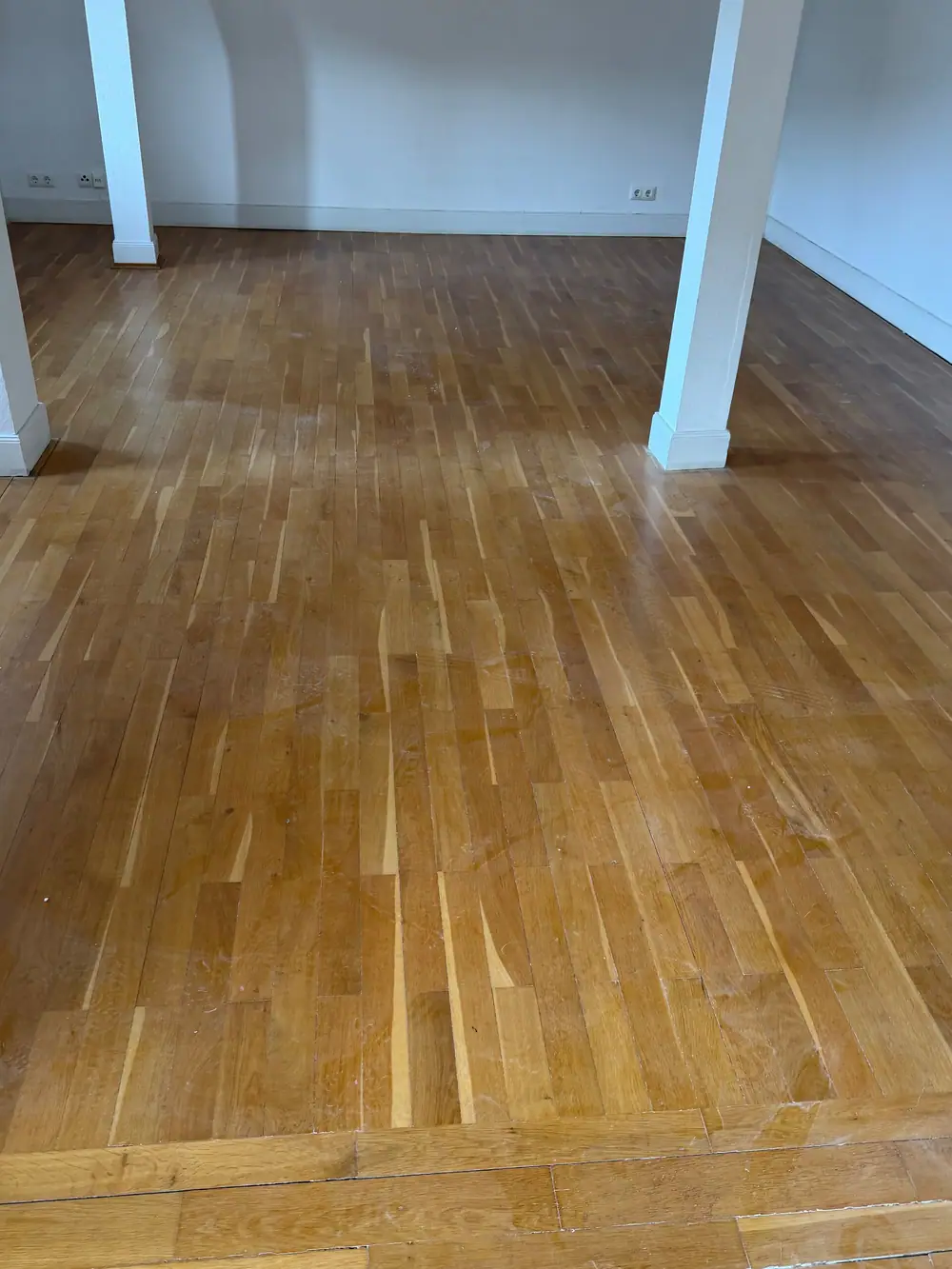 Leerer Raum mit weiß gestrichenen Stützen, Sockelleisten und einem Holzfußboden aus schmalen Stäben.