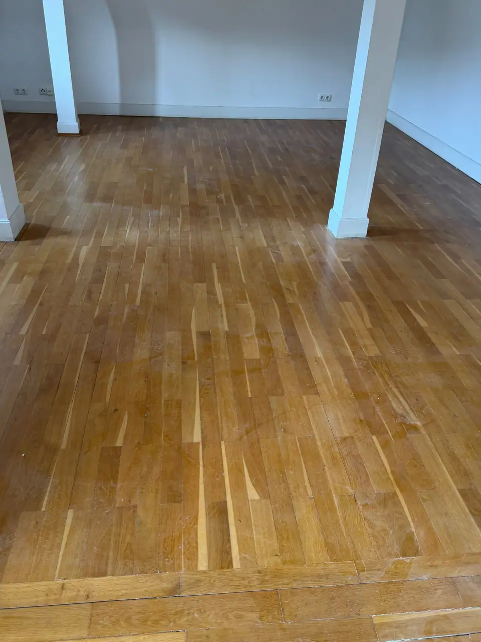 Leerer Raum mit weiß gestrichenen Stützen, Sockelleisten und einem Holzfußboden aus schmalen Stäben.