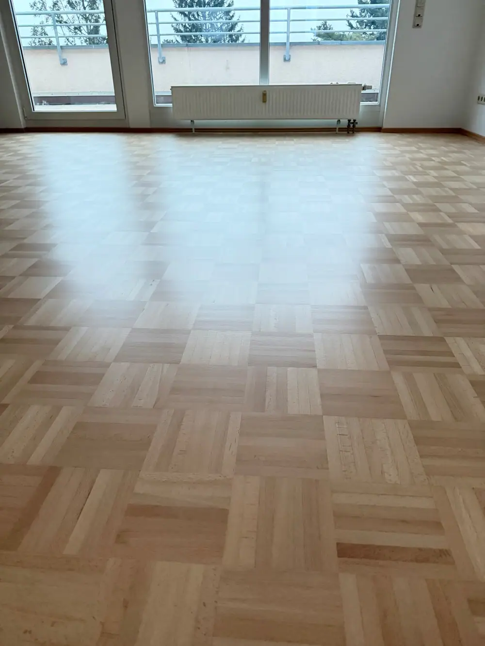 Ansicht der hellen Bodenfläche mit quadratisch angeordneten Mosaikparkettfeldern aus Buche im Bereich vor Fenster und Heizkörper.