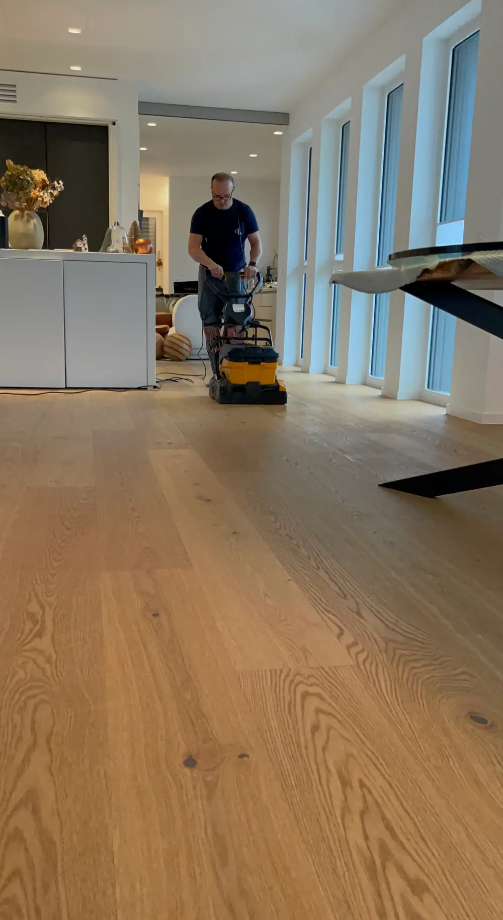 Mitarbeiter arbeitet mit einer Bodenreinigungsmaschine auf einem hellen Holzboden entlang einer Reihe hoher Fenster.