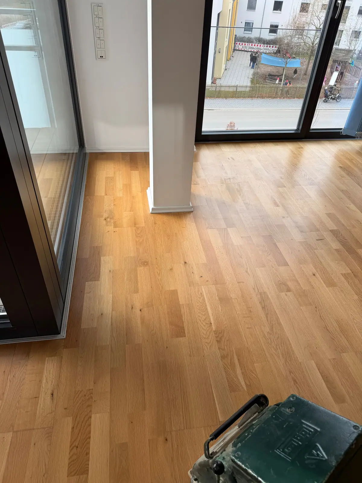 Eiche-Parkett neben einer großen Fensterfront; rechts unten ist eine Schleifmaschine zu sehen.