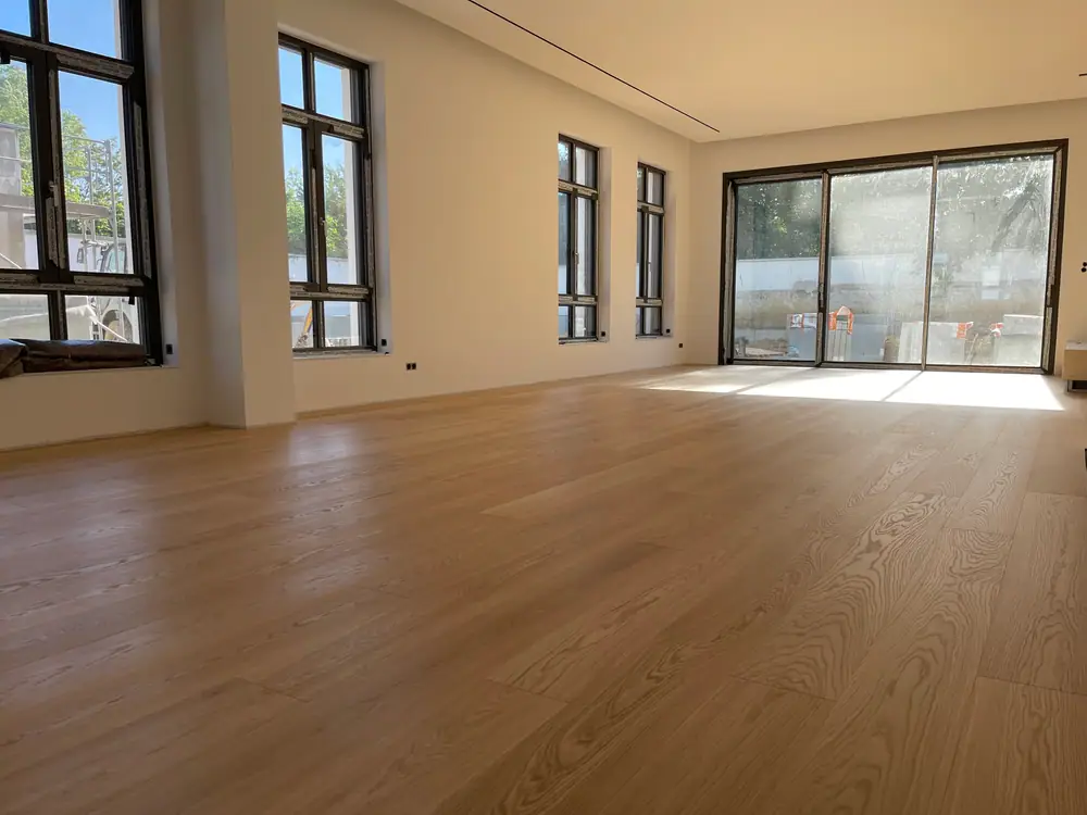 Großzügiger Innenraum mit breiten hellen Holzdielen und hohen, noch abgeklebt wirkenden Fenstern.