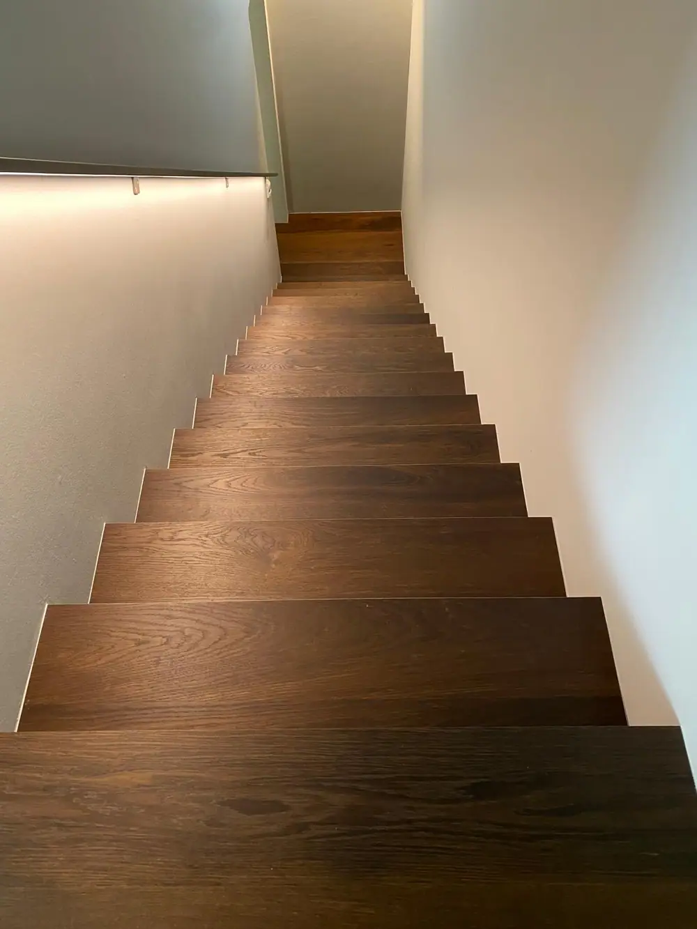 Blick von oben auf einen geraden Treppenlauf mit dunkelbraunen Holzstufen und heller Wand auf beiden Seiten.