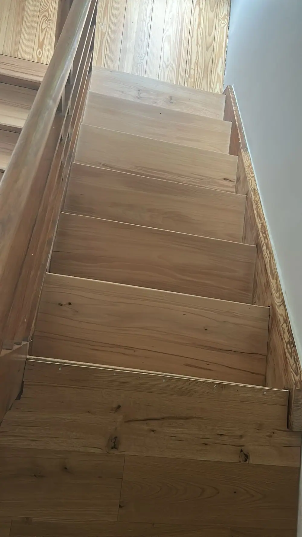 Blick von unten auf eine Holztreppe mit breiten Stufen, Holzgeländer und seitlicher Wandleiste.