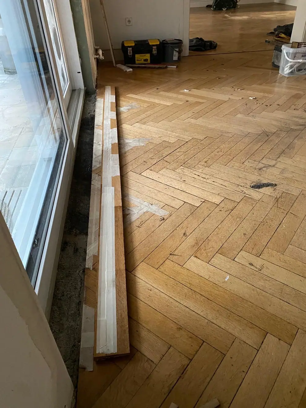 Fischgrätparkett mit einem geöffneten Anschlussbereich entlang eines großen Fensters.