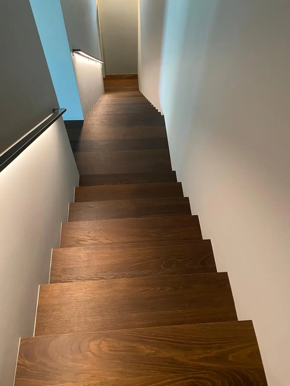 Blick von oben über dunkelbraune Holzstufen in einem schmalen Treppenhaus mit hellen Wänden und Handlauf.