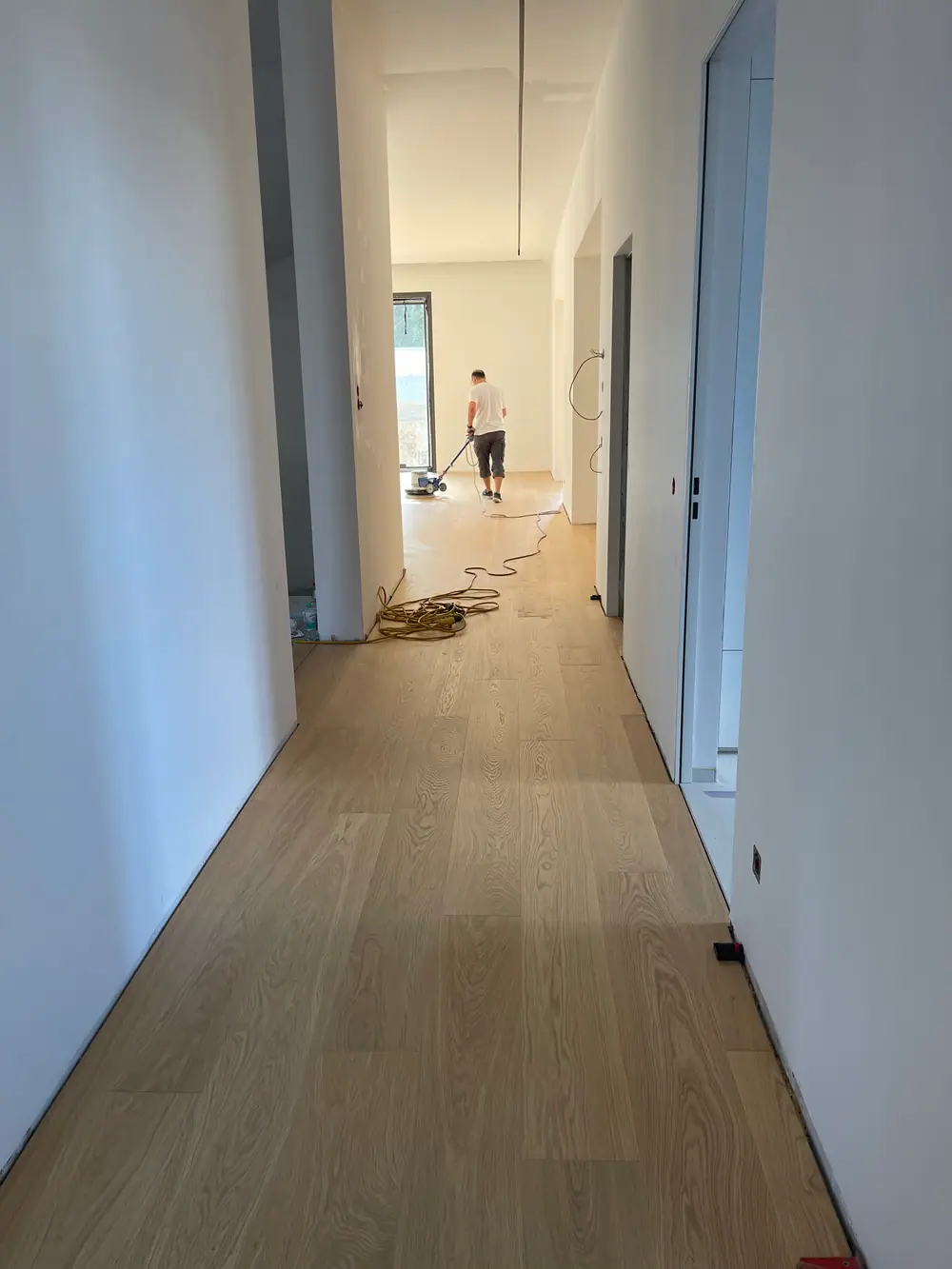Langer Flur mit hellem Dielenboden, weißen Wänden, Kabeln und einer Person mit Bodenmaschine.
