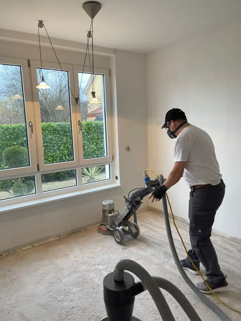 Mitarbeiter arbeitet mit einer Estrichfräse auf dem Boden vor einer großen Fensterfront in einem hellen Innenraum.
