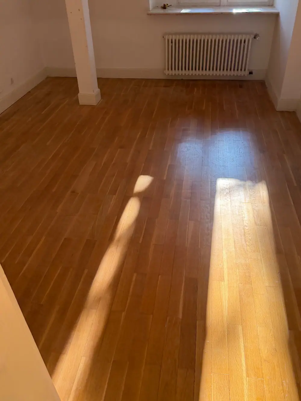 Leerer Raum mit weißer Wand, Heizkörper und einem Holzfußboden, auf den Sonnenlicht durch ein Fenster fällt.