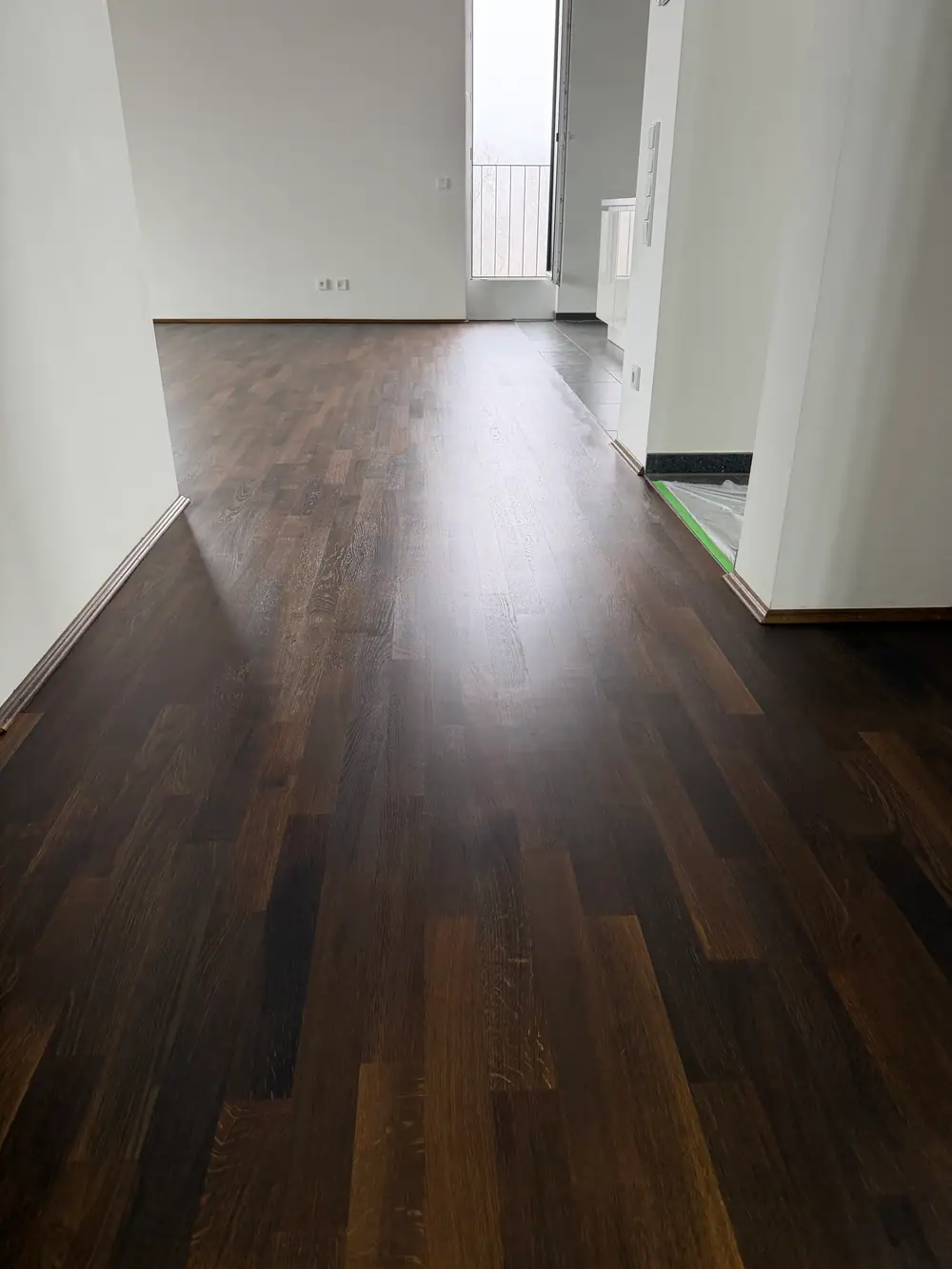 Dunkler Holzboden in einem Flur mit weißen Wänden und geöffneten Türen.