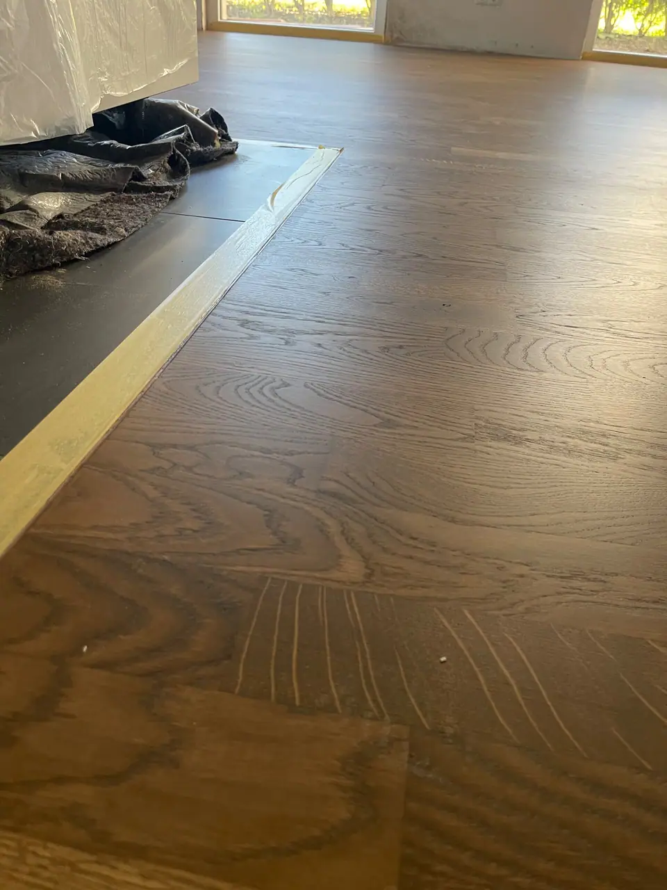 Dunkles Parkett mit sichtbarer Holzmaserung an einem mit Klebeband abgegrenzten Übergang zu einem dunklen Bodenbereich.