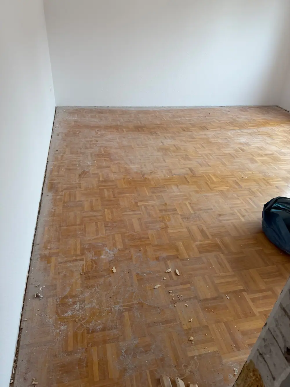 Leerer Raum mit älterem Mosaikparkett aus Eiche, dessen Oberfläche Staub, Gebrauchsspuren und ungleichmäßige Bereiche zeigt.