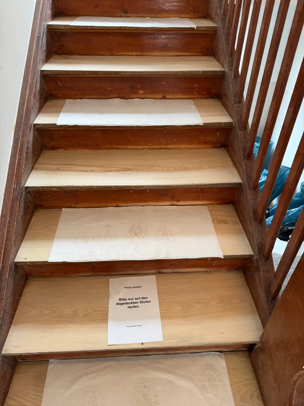 Holztreppe während der Arbeiten; mehrere helle Stufen sind mit Papier abgedeckt, auf einer Stufe liegt ein Hinweis zur frischen Lackierung.