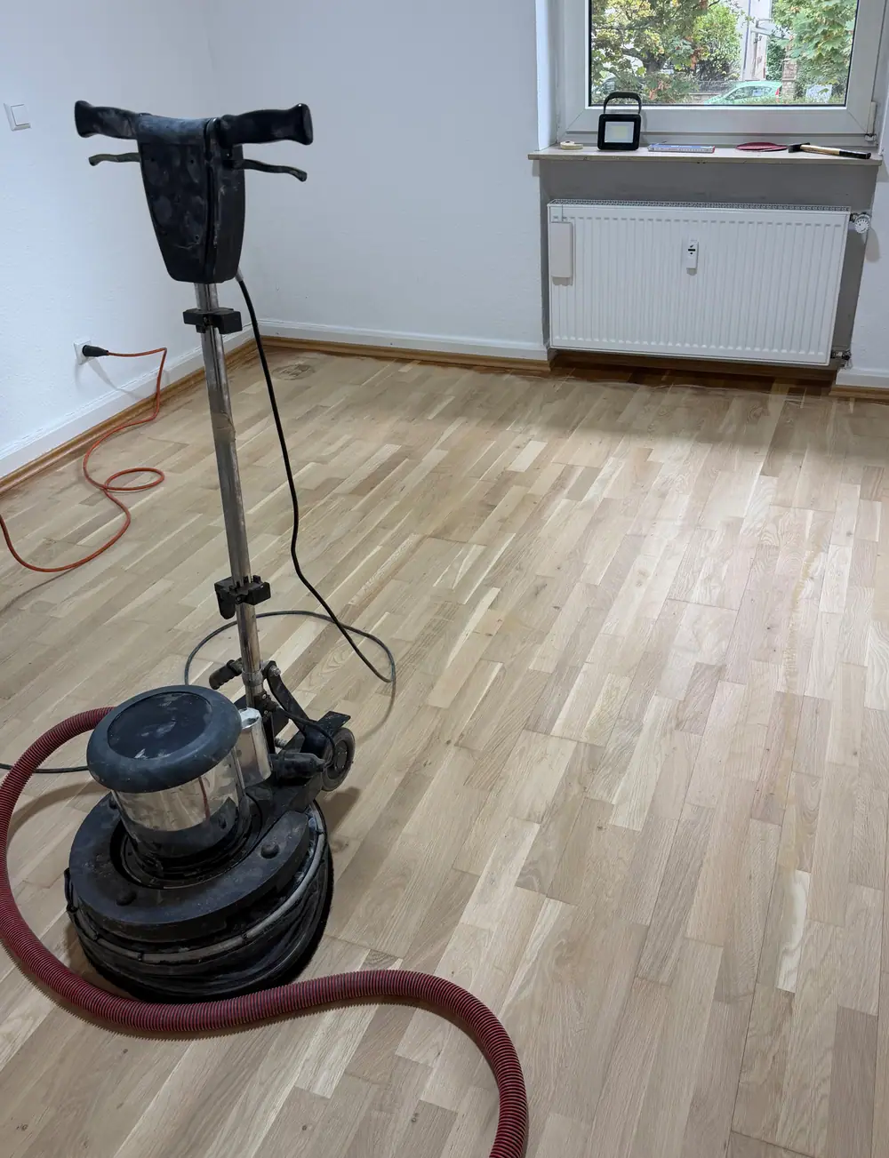 Helle, angeschliffene Fischgrätparkettfläche mit Bodenschleifmaschine und rotem Absaugschlauch.