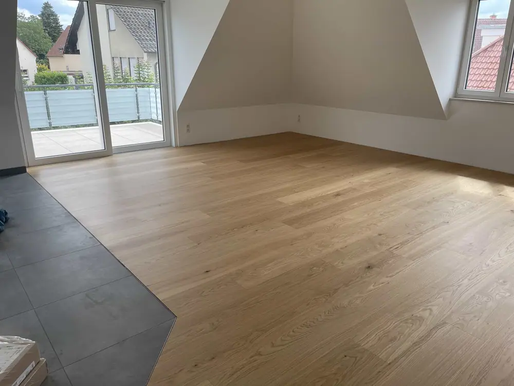Heller Holzboden in einem offenen Raum mit großformatigen grauen Fliesen am seitlichen Bodenübergang, Fenstern und Balkontür.
