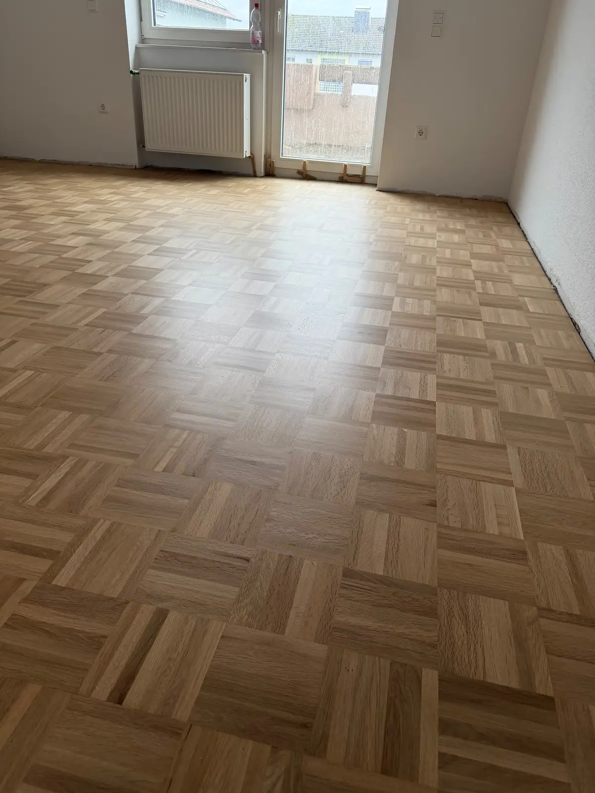 Weite Raumansicht des sanierten Eiche-Mosaikparketts mit hellem Würfelmuster, Fenster, Balkontür und Heizkörper.