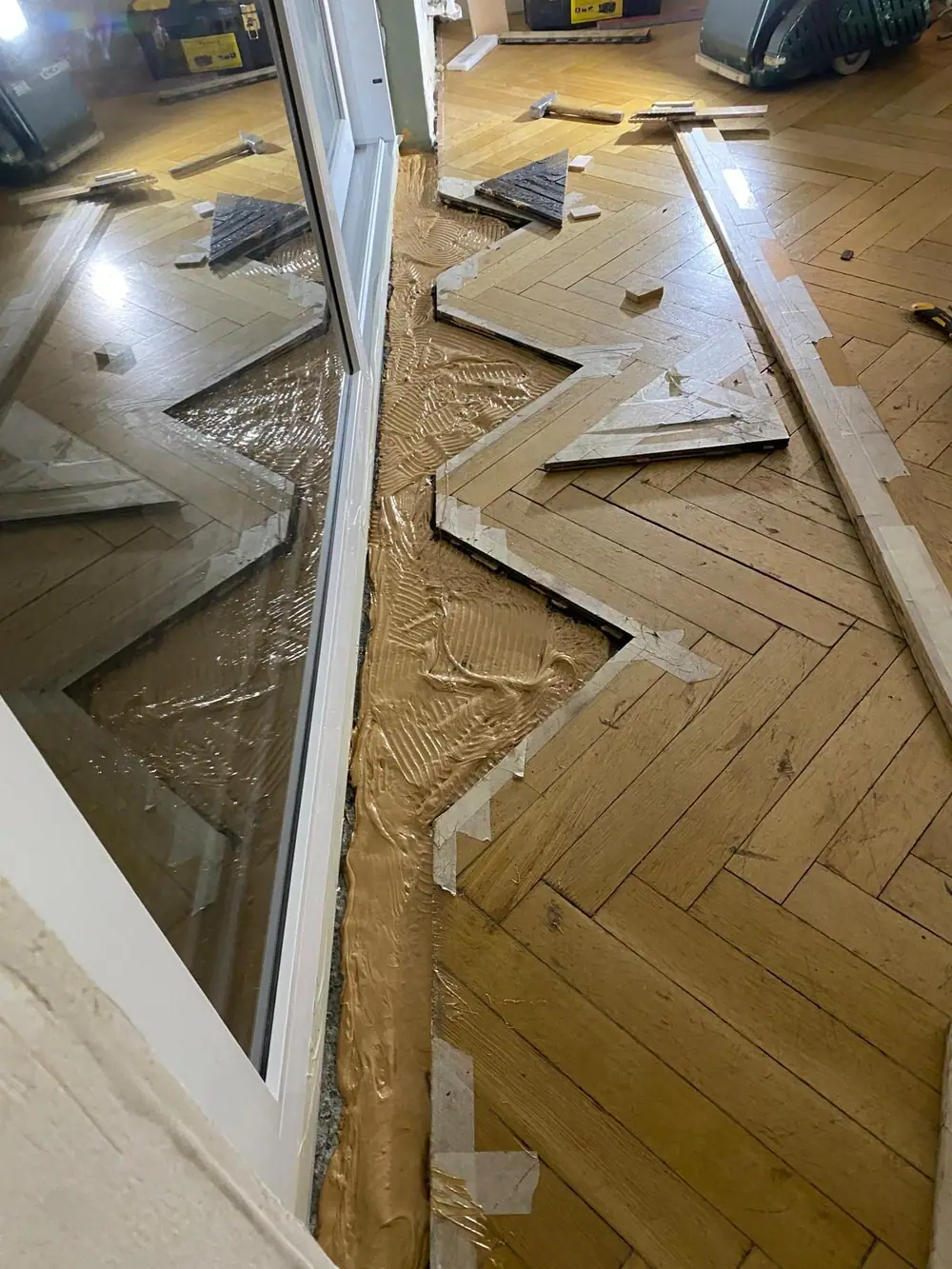 Arbeitsbereich am Fenster mit Kleber auf dem freigelegten Untergrund und Parketteilen.