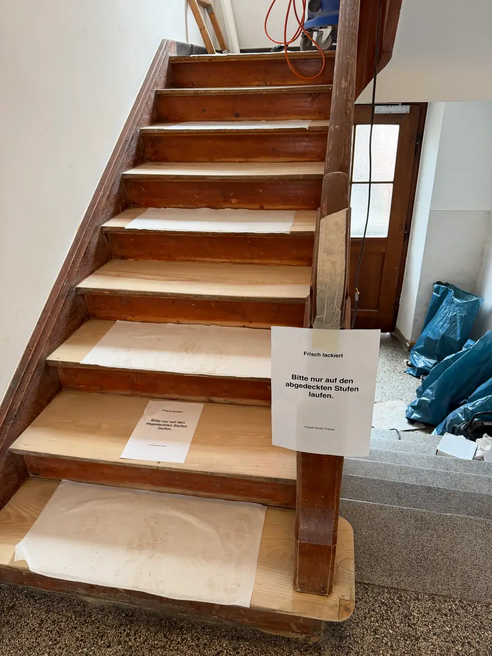 Teilansicht einer Holztreppe mit Papierabdeckungen und Hinweisschildern zur frischen Lackierung.