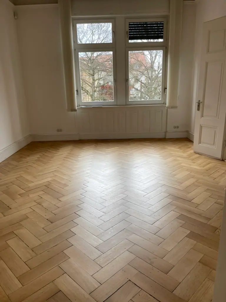 Heller Raum mit Eiche Stabparkett im Fischgrätmuster, großem Fenster, weißer Wandvertäfelung und geöffneter Kassettentür.