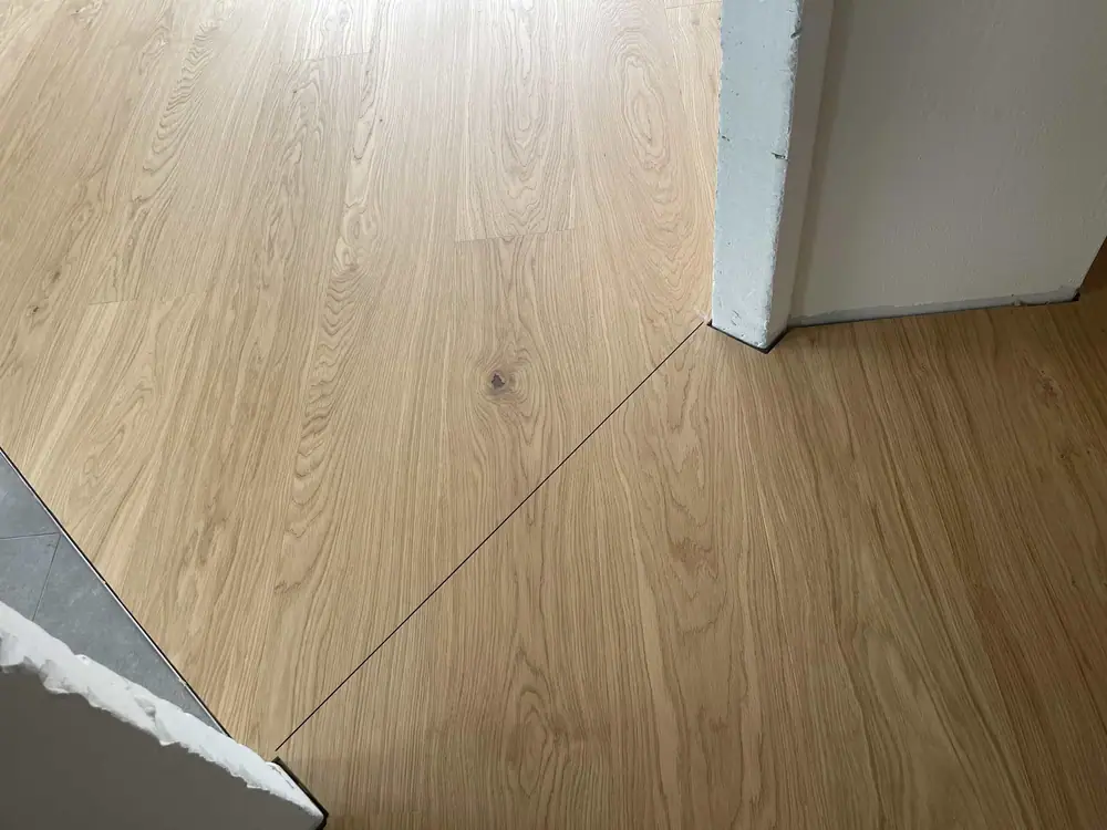 Heller Holzboden mit zugeschnittener Kante an einem weißen Wandvorsprung und angrenzendem Fliesenbereich.