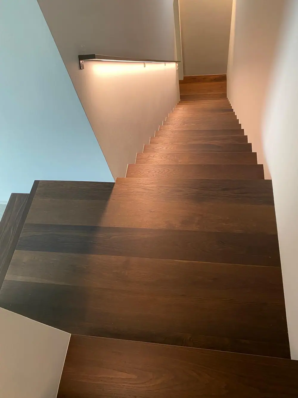 Blick über ein dunkelbraunes Holzpodest auf einen absteigenden Treppenlauf mit hellem Wandbereich und Handlauf.