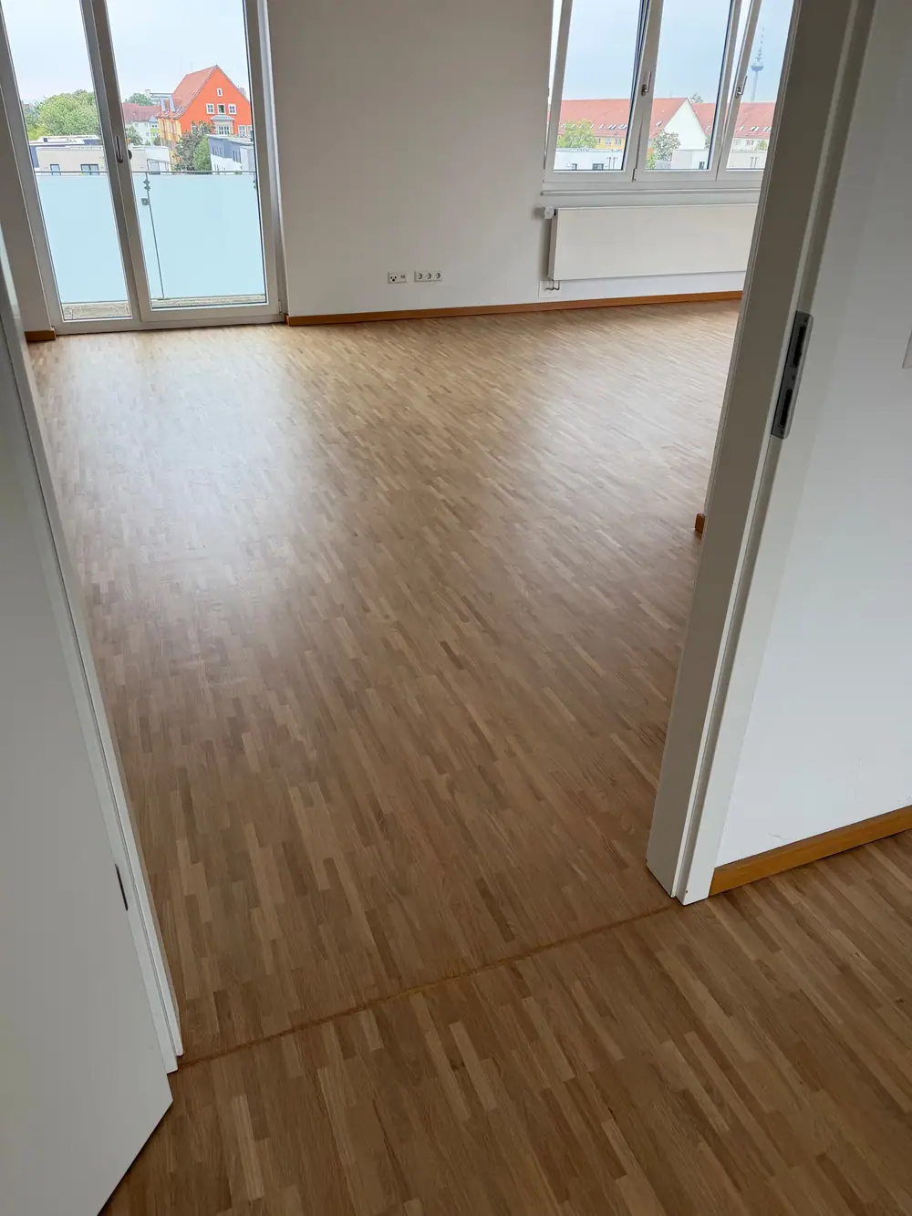 Blick durch eine Tür in einen hellen Raum mit geschliffenem Eichenparkett, Balkonfenster und Heizkörper.