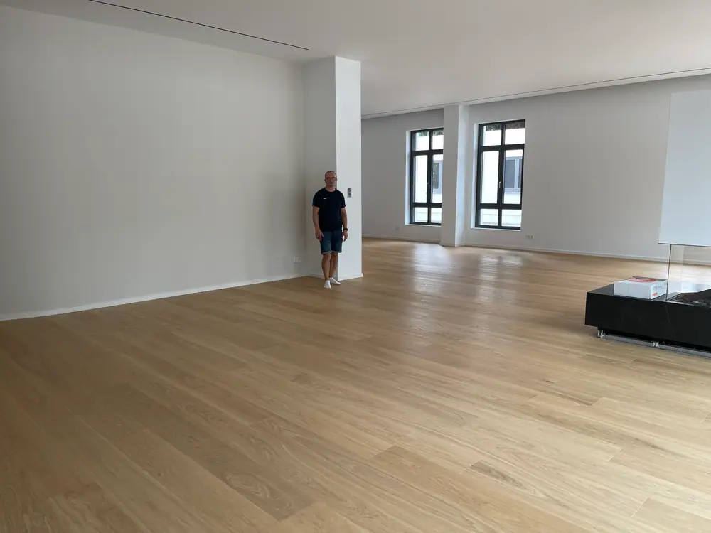 Weiter Innenraum mit hellem Holzdielenboden, weißen Wänden, hohen Fenstern und dunklem Sockelbereich.