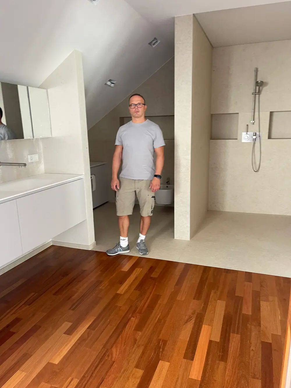 Badezimmer mit Doussie-Parkett, hellem Fliesenbereich, Dusche und einer stehenden Person.