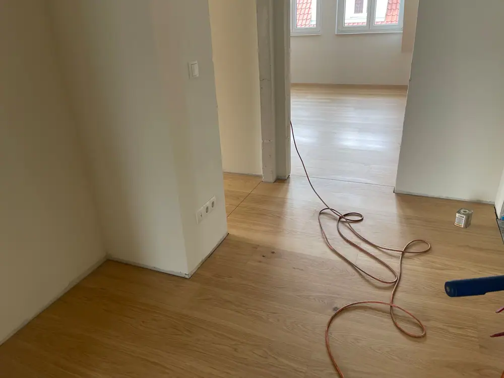 Heller Holzboden in einem Durchgang mit weiß verputzten Wänden und sichtbarem Arbeitskabel auf dem Boden.