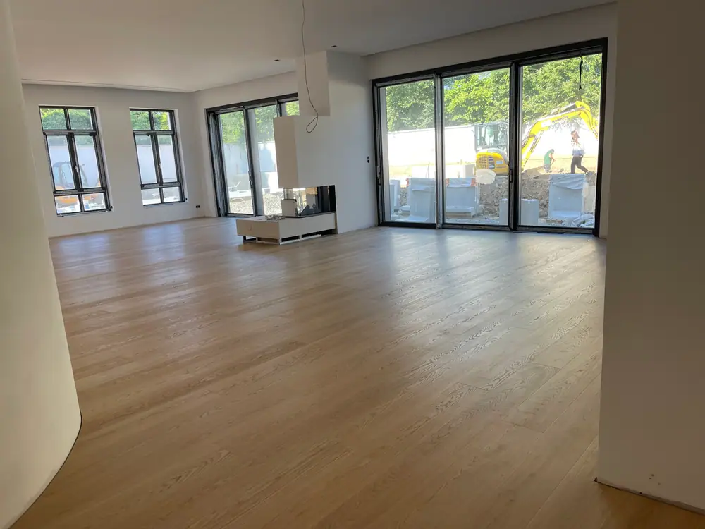 Großer unfertiger Wohnraum mit hellem Dielenboden, hohen Fensterflächen und mittigem Kaminelement.
