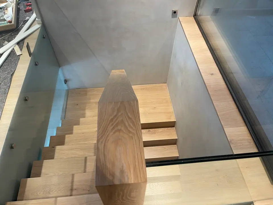 Blick von oben in ein Treppenhaus mit Holzstufen, Glasgeländer und dunklem Fensterrahmen.