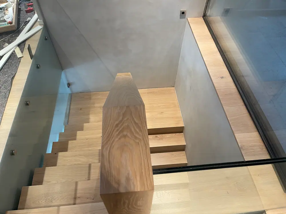 Blick von oben in ein Treppenhaus mit Holzstufen, Glasgeländer und dunklem Fensterrahmen.