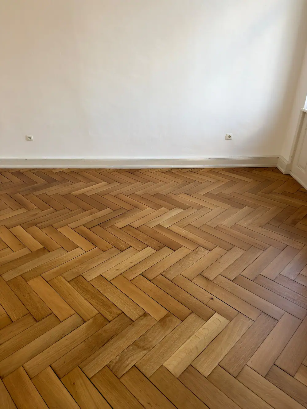Fischgrätparkett vor einer weißen Wand mit Sockelleiste und Steckdosen.
