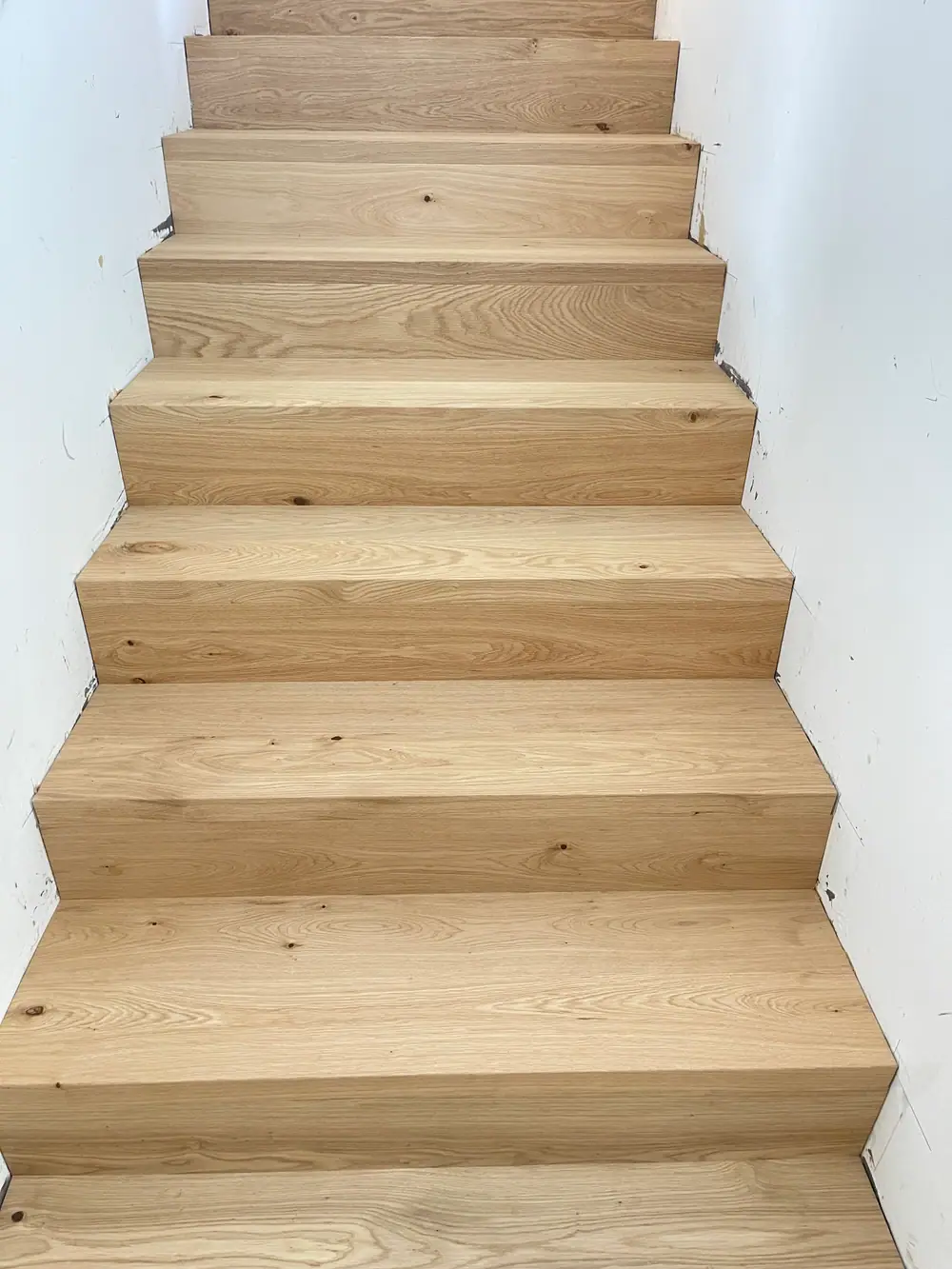 Gerade Treppe mit breiten Holzstufen, sichtbaren Aststellen und weißen Wänden.