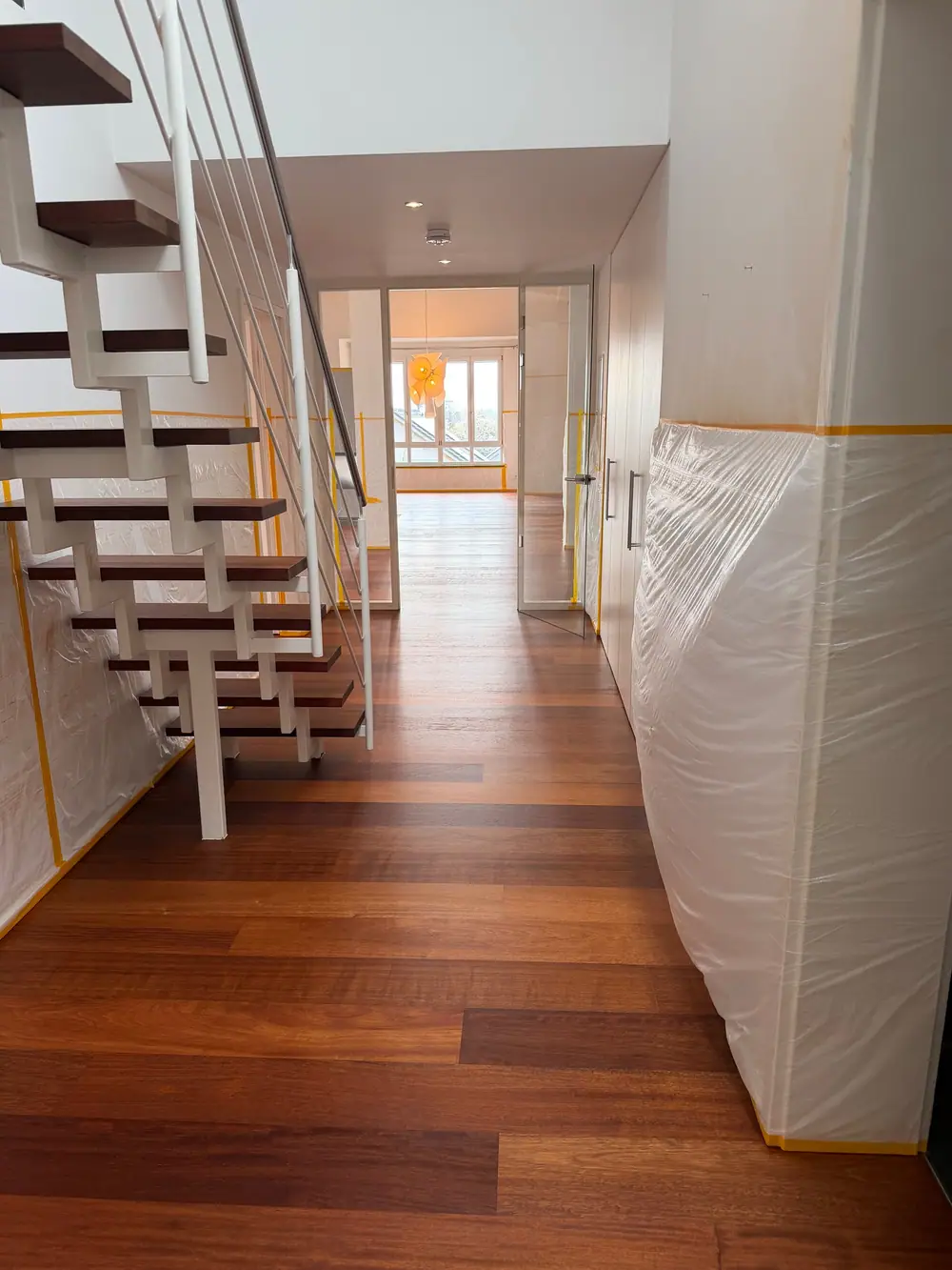 Flur mit dunklem Dielenboden, offener weißer Treppe, abgedeckten Wänden und Blick in den Wohnraum.