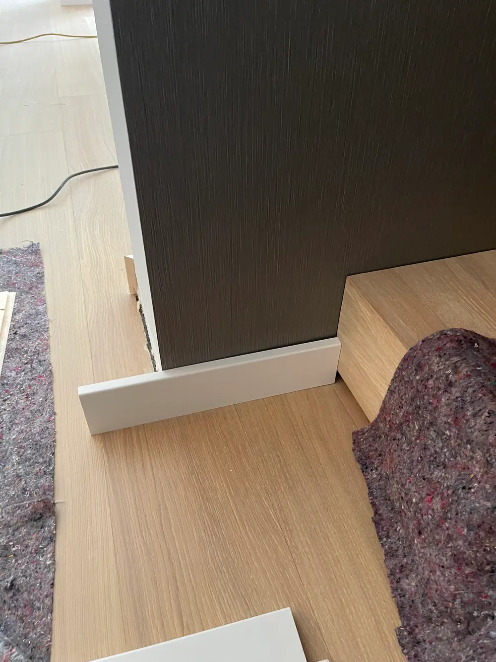 Helle Sockelleiste vor einem dunklen Möbelteil auf einer hellen Holzdielenfläche.