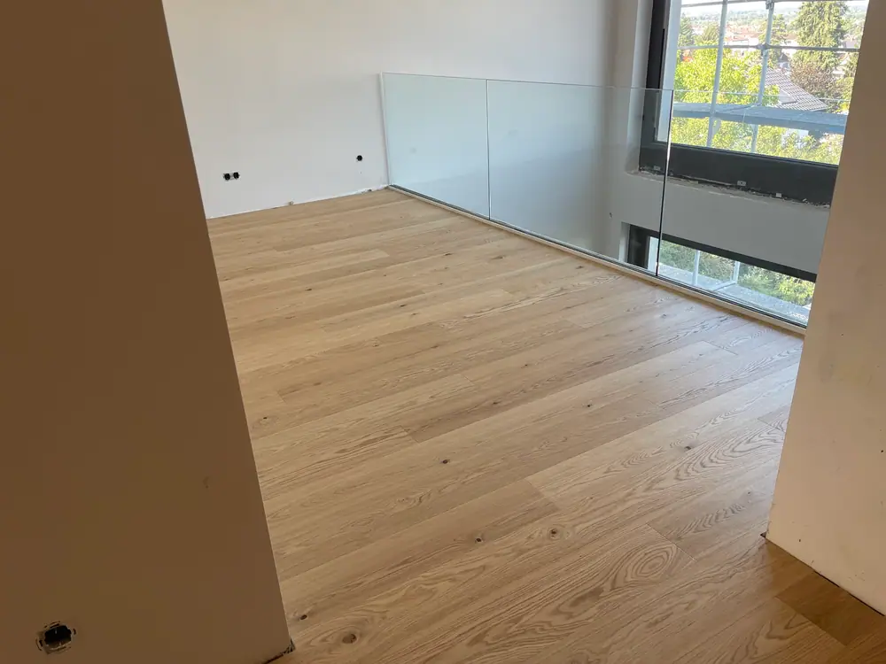 Heller Dielenboden auf einer Galerie mit Glasgeländer, großen Fensterflächen und unfertigen Wandanschlüssen.