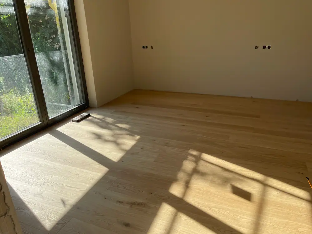 Heller Dielenboden mit sichtbarer Maserung und Sonnenlicht vor einer hohen Fensterfront.